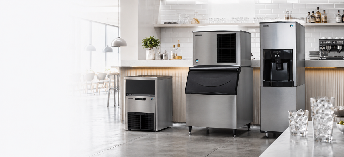 Three stainless steel commercial ice machines in different sizes arranged in a bright, modern kitchen setting with white tile walls, wood countertops, and glassware on the counter.